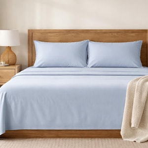 May include: A light blue bed sheet set on a wooden bed frame. The bed is made with two matching pillowcases and a folded beige blanket. A wooden nightstand with a lamp and a potted plant are on the left.