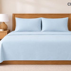Op de afbeelding: Een bed met een lichtblauwe sprei en twee bijpassende lichtblauwe kussenslopen. Het bed heeft een houten hoofdeinde en frame. De tekst Cloud Blue is zichtbaar in de rechterbovenhoek van de afbeelding en geeft de kleur van de beddengoedset aan.