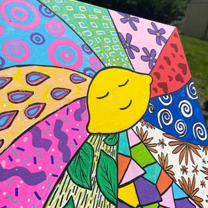 May include: A colorful canvas painting with a smiling yellow lemon in the center. The lemon is surrounded by different patterns and designs, including circles, flowers, leaves, and geometric shapes. The painting is done in a variety of bright colors, including pink, blue, green, yellow, orange, and red.