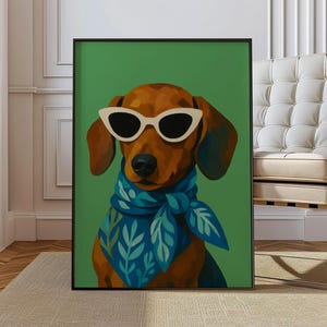 May include: A framed art print featuring a dachshund wearing white sunglasses and a blue scarf with a leaf pattern. The dog is set against a green background. The artwork is displayed in a room with a beige rug and a white chair.