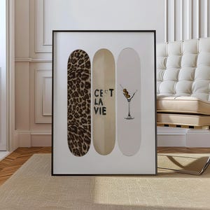 May include: Framed art print featuring three skateboard decks. One deck has a leopard print, the second has the text "C'est La Vie", and the third has a cocktail illustration. The frame is black, and the artwork is displayed on a beige rug.