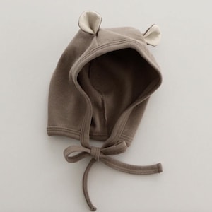 Puede incluir: Un gorro de bebé marrón con capucha y orejas de oso adjuntas. El gorro tiene un cierre de lazo debajo de la barbilla. El interior de las orejas es de color beige claro. El gorro está hecho de una tela de punto suave.