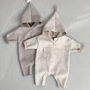 May include: Two baby rompers in light beige and taupe, each featuring a pointed hood with a small pom-pom at the tip, short sleeves with folded cuffs, a front pocket with a flap, and snap closures along the inner leg for easy access.