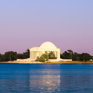 May include: A scenic view of the Jefferson Memorial in Washington, D.C., with its white marble structure and domed roof. The memorial is reflected in the blue water of the Tidal Basin, under a purple and blue sky.