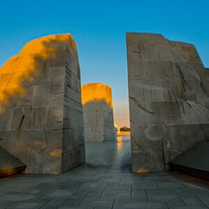 May include: Exterior view of the Martin Luther King, Jr. Memorial in Washington, D.C. The memorial features large, light gray granite structures with golden sunlight illuminating the tops. The sky is a clear blue.