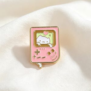 May include: A pink enamel pin in the shape of a retro handheld game console. The screen displays a white cat with a heart and star. The pin has gold-colored trim and button details.