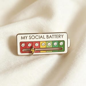Könnte beinhalten: Eine rechteckige Emaille-Anstecknadel mit dem Text "MY SOCIAL BATTERY" oben. Die Anstecknadel zeigt eine Batteriestandsanzeige mit einer Reihe von Emoticons, die von traurig bis glücklich reichen, und einen Blitz. Die Batteriestandsanzeige ist rot, gelb und grün gefärbt.