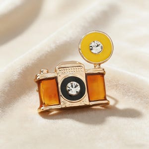 Puede incluir: Broche de cámara dorada con detalles ámbar y flash amarillo. La lente de la cámara es negra con un cristal central, y el flash tiene un cristal grande. Esta joya es un accesorio con temática de fotografía.