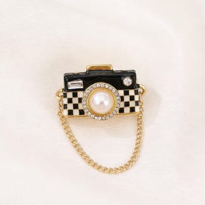Puede incluir: Broche de cámara en blanco y negro con cadena dorada. La cámara tiene una perla en el centro, rodeada de cristales transparentes. El cuerpo de la cámara es negro con un patrón de cuadros en la parte inferior.