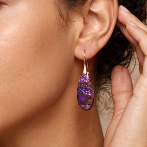 May include: A teardrop-shaped earring featuring a polished purple and dark-toned stone with natural veining, suspended from a thin gold-colored metal hook. The stone displays a mottled pattern of deep violet, black, and metallic copper-colored flecks.