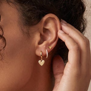 May include: A set of gold-toned earrings consisting of a small heart-shaped stud, a thin hoop earring with embedded clear stones, and a small hoop earring featuring a dangling heart-shaped charm, all worn in a single earlobe.