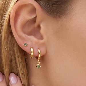 May include: A set of gold-toned jewelry consisting of a small stud earring with a green gemstone and two gold hoop earrings. One hoop is plain, while the other features a small, dangling green gemstone charm that matches the stud earring.