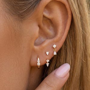 May include: A set of four gold-toned earrings worn in a single ear. The collection includes a small hoop with three embedded stones, a tiny triangular stud, a small vertical bar stud with two stones, and a drop earring featuring a single stone.
