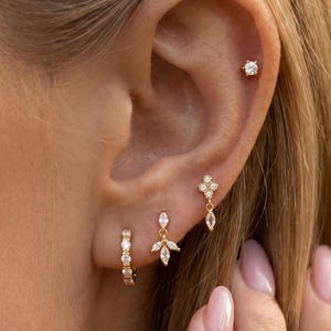 May include: A set of gold-toned earrings featuring a small hoop with embedded clear stones, a dangling charm with three marquise-cut stones, a four-stone clover-shaped stud, and a single round stone stud worn in the upper ear cartilage.