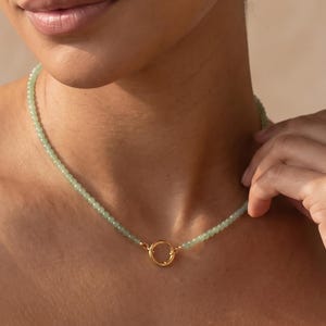 May include: A beaded necklace featuring small, round, light green stones strung together. The necklace is secured with a circular gold-toned metal clasp positioned at the front, connecting the two ends of the beaded strand in a simple, clean design.