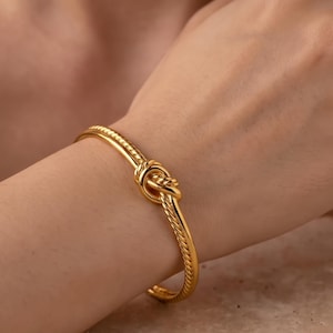 May include: A gold-toned metal bangle bracelet featuring a twisted rope texture and a central knot design. The bracelet has a polished finish and a circular shape that rests on the wrist, showcasing a continuous twisted pattern throughout the band.