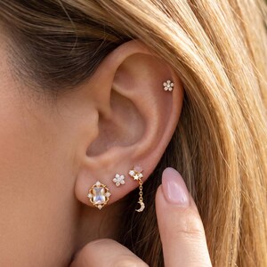 May include: A collection of gold-toned earrings, including a flower stud, a square-shaped earring with a gemstone, a cluster of flowers, and a dangling moon and star design. The earrings are displayed on an ear, showcasing their delicate details and varied styles.