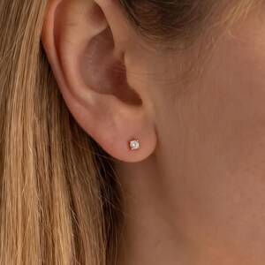 Peut inclure: Une petite boucle d'oreille à clou ornée d'une pierre transparente ronde, fixée dans une monture métallique à griffes et portée au lobe de l'oreille. La pierre facettée reflète la lumière, créant un éclat brillant sur la peau.