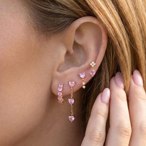 May include: A collection of pink gemstone earrings in various styles. The earrings include small hoops, dangling heart-shaped stones, and a stud with a flower design. The jewelry is set against a blurred background, highlighting the earrings' details.