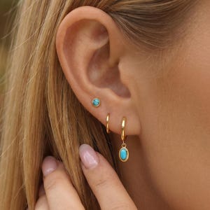 May include: A set of gold-toned earrings consisting of a small stud with a round turquoise stone, a plain gold hoop, and a gold hoop featuring a dangling oval turquoise charm. The jewelry is worn in three separate piercings along the earlobe.