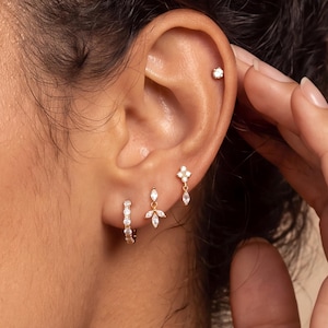 May include: A set of three gold earrings worn in the earlobe and cartilage. The lowest earring is a small hoop with embedded stones, the middle is a dangling marquise-cut stone charm, and the top is a small stud with a single clear stone.