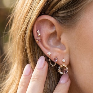 May include: Close-up of multiple gold earrings with small gemstones and floral designs. One earring features a chain connecting two floral studs. Another earring has a circular design with small crystals and a gemstone in the center.