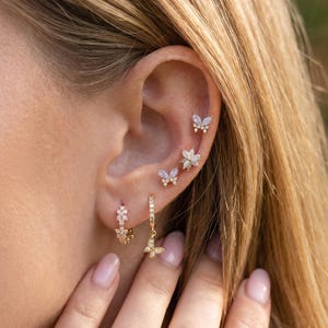 May include: A collection of gold-toned earrings featuring butterfly and floral designs. The earrings include small studs and dangling hoops, all adorned with sparkling gemstones. The image showcases multiple ear piercings with the jewelry.