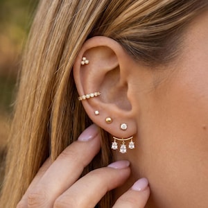 May include: An ear displaying a collection of gold earrings. There's a helix earring, a small stud, a gold ball, a diamond stud, and a distinctive earring with three hanging teardrop gems. The skin tone is fair, and the background is out of focus.