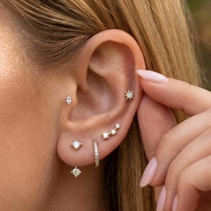 May include: Close-up of an ear adorned with multiple gold earrings. The earrings include a starburst stud, a small hoop, a constellation design, and various gemstone studs. The ear is set against a blurred background.
