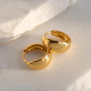 Puede incluir: Un par de pendientes de aro dorados con una superficie lisa, pulida y redondeada. Los pendientes presentan un diseño grueso y macizo con forma circular y un mecanismo de cierre de bisagra, reflejando la luz en su acabado metálico dorado.
