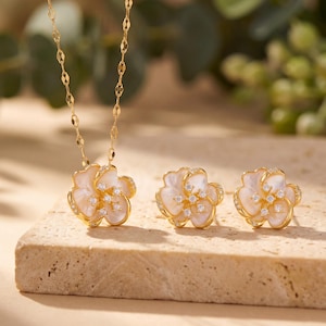 May include: A gold-toned necklace and matching earrings set. The jewelry features a flower design with white petals, small clear stones, and a gold-colored setting. The necklace and earrings are displayed on a light-colored surface.