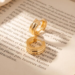 May include: A pair of small gold hoop earrings featuring a triple-band design with a polished finish. The earrings are circular with a hinged closure mechanism, showcasing a layered, ribbed texture that creates a distinct, multi-dimensional look.