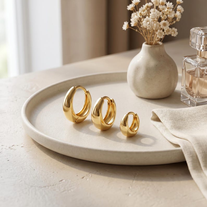 May include: A set of three gold-toned hoop earrings in graduated sizes, featuring a smooth, polished finish and a hinged closure. The earrings are arranged in a row from largest to smallest, showcasing a rounded, thick profile and a reflective surface.