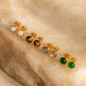May include: Four pairs of stud earrings with gold-toned metal settings. Each pair features a different round gemstone: iridescent white opal, brown tiger's eye, grey labradorite, and solid green stone. The earrings are arranged in a diagonal row.
