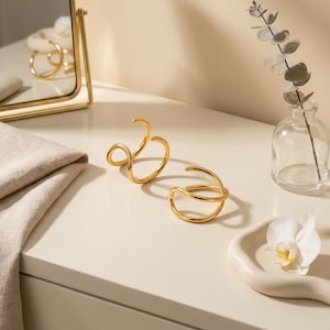May include: Two gold-toned metal ear cuffs with a smooth, polished finish. Each piece features a double-loop design with an open, adjustable structure. The cuffs are crafted from a continuous, rounded wire that curves to form two overlapping circles.