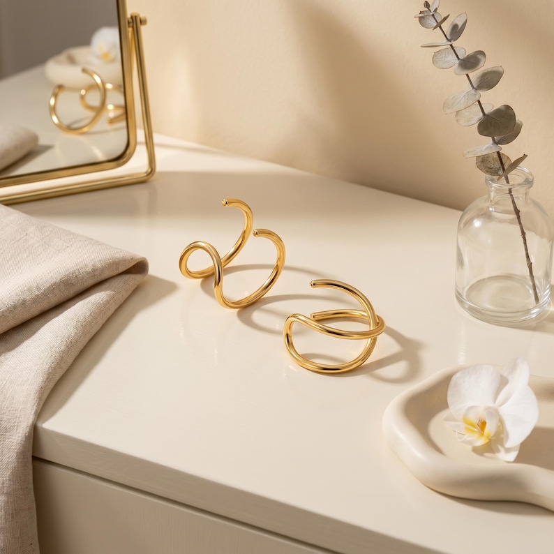 May include: Two gold-toned metal ear cuffs with a smooth, polished finish. Each piece features a double-loop design with an open, adjustable structure. The cuffs are crafted from a continuous, rounded wire that curves to form two overlapping circles.