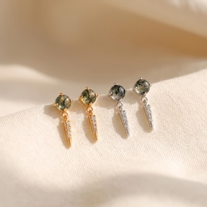 May include: Two pairs of drop earrings featuring round moss agate stones set in prongs. One pair is gold-toned and the other is silver-toned. Each earring has a dangling, slender, triangular charm encrusted with small, clear, sparkling stones.