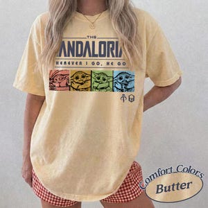 May include: Butter-colored t-shirt featuring the text "THE MANDALORIAN" above the phrase "WHEREVER I GO, HE GO." The shirt displays four colorful images of Grogu. The shirt is a Comfort Colors brand.