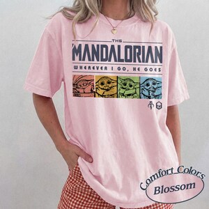 May include: A light pink t-shirt featuring the text "THE MANDALORIAN" above the phrase "WHEREVER I GO, HE GOES." Below the text are four images of Grogu in different colors. The shirt also has the "Comfort Colors Blossom" logo.