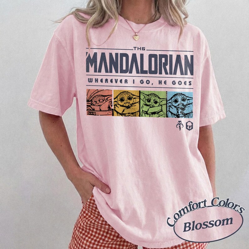 May include: A light pink t-shirt featuring the text "THE MANDALORIAN" above the phrase "WHEREVER I GO, HE GOES." Below the text are four images of Grogu in different colors. The shirt also has the "Comfort Colors Blossom" logo.