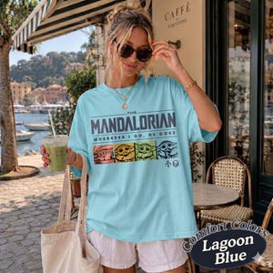May include: A light blue t-shirt with the text "THE MANDALORIAN" and "WHEREVER I GO, HE GOES" in dark blue. Below the text are four images of Grogu in different colors. The shirt is labeled "Comfort Colors Lagoon Blue".