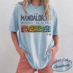 May include: Light blue t-shirt with the text "THE MANDALORIAN" and "WHEREVER I GO, HE GOES." The shirt features four colorful images of Grogu. The shirt is a Comfort Colors Chambray.