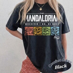 May include: Black t-shirt featuring the text "THE MANDALORIAN" above the phrase "WHEREVER I GO, HE GOES." The shirt includes a graphic of the character in four different color blocks. The words "Comfort Colors Black" are printed on the bottom right.