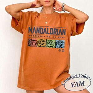 May include: An orange Comfort Colors t-shirt featuring the text "THE MANDALORIAN" above a colorful graphic of Grogu. The phrase "WHEREVER I GO, HE GOES" is printed below the title. The shirt has a relaxed fit.
