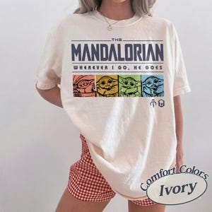 May include: Ivory-colored t-shirt featuring the text "THE MANDALORIAN" above the phrase "WHEREVER I GO, HE GOES." Below the text are four colorful images of Grogu. The shirt is a Comfort Colors brand.