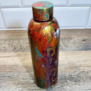 Può includere: Una bottiglia decorativa dipinta a mano con coperchio abbinato. La bottiglia presenta un vivace design astratto con tonalità rosse, arancioni, oro, verde acqua e viola. La bottiglia è alta circa 20 cm.