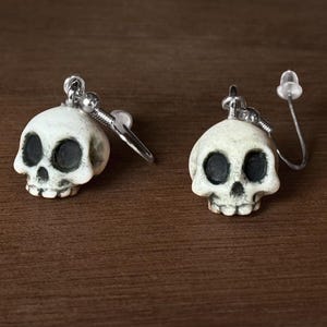 May include: A pair of silver skull earrings with white enamel. The skulls have black eye sockets and a detailed nose and mouth.