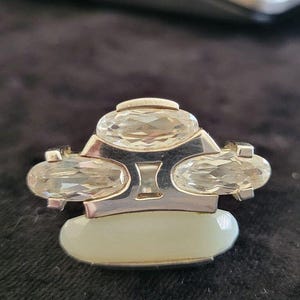 May include: A silver-toned ring featuring three faceted, clear, oval-shaped stones arranged horizontally across the top, with a single, smooth, opaque, pale white oval stone set horizontally at the base of the metal band structure.