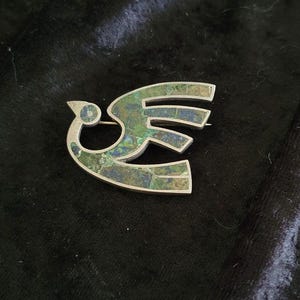 May include: A silver-toned brooch shaped like a stylised bird in flight. The body of the bird is inlaid with a mosaic of small, flat, irregular pieces of stone in shades of deep forest green, moss green, and dark blue, set within a metal frame.