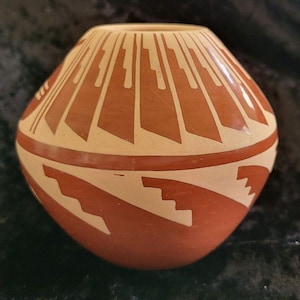 May include: A rounded terracotta-colored ceramic vase featuring a wide opening and a tapered base. The surface is decorated with cream-colored geometric patterns, including vertical rays around the neck and stepped designs around the body.